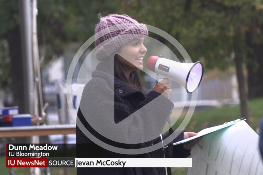 A woman in Dunn Meadow speaking into a megaphone. Lower thirds: Dunn Meadow. IU Bloomington. IU NewsNet source: Jevan McCosky.